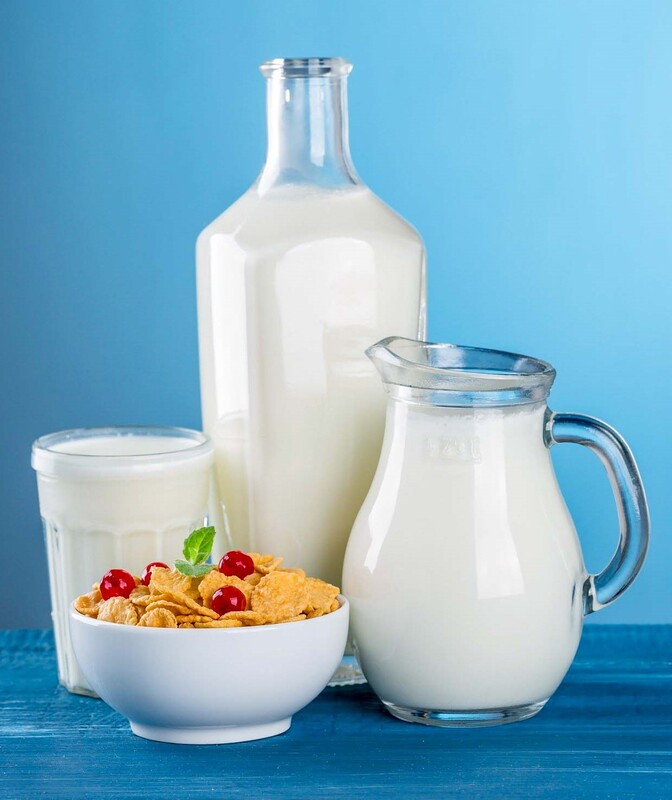 A glass, a bottle, and a pitcher of milk on a blue table around a white bowl of cornflakes